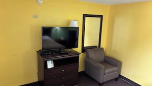 Une chambre d’hôtel avec un téléviseur à écran plat posé sur une commode, un fauteuil gris et un grand miroir au mur.