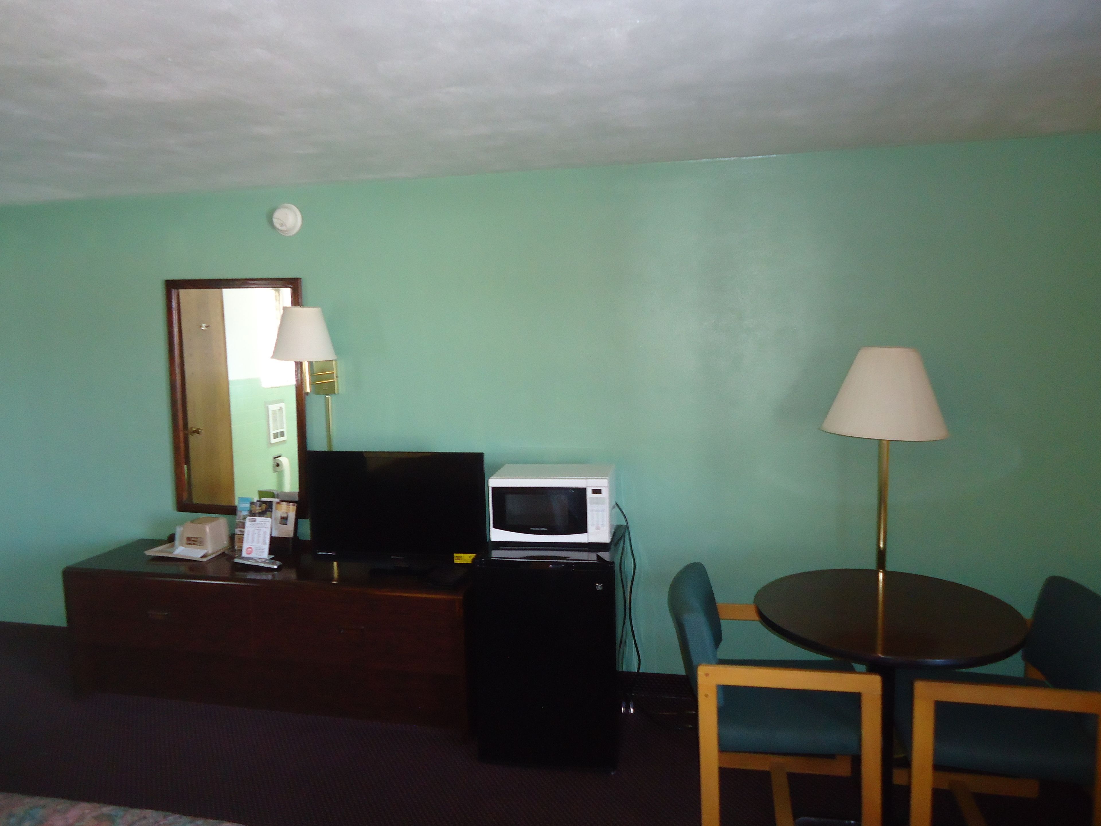 Una habitación con paredes color turquesa, un escritorio de madera con un televisor, un microondas y una lámpara. Hay una mesa redonda con dos sillas y un pequeño armario con un espejo.