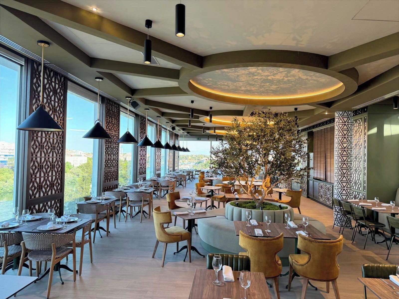Ein Restaurant mit einer zentralen Sitzgruppe, die einen Baum und eine runde Leuchte beinhaltet. Tische sind um die Sitzgruppe angeordnet, und große Fenster bieten einen Blick ins Freie.