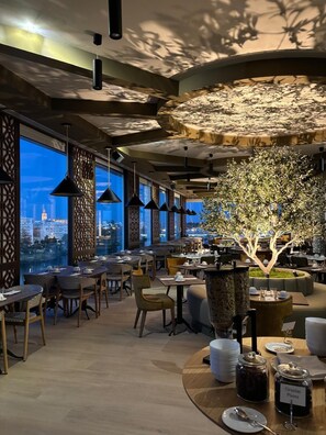A restaurant with a view of a city skyline, featuring a large tree centerpiece and a variety of seating arrangements.