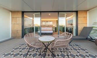 Un balcon doté de meubles en osier, d’un sol à motifs et d’une chambre à coucher avec un lit et des rideaux visibles à travers des portes vitrées.