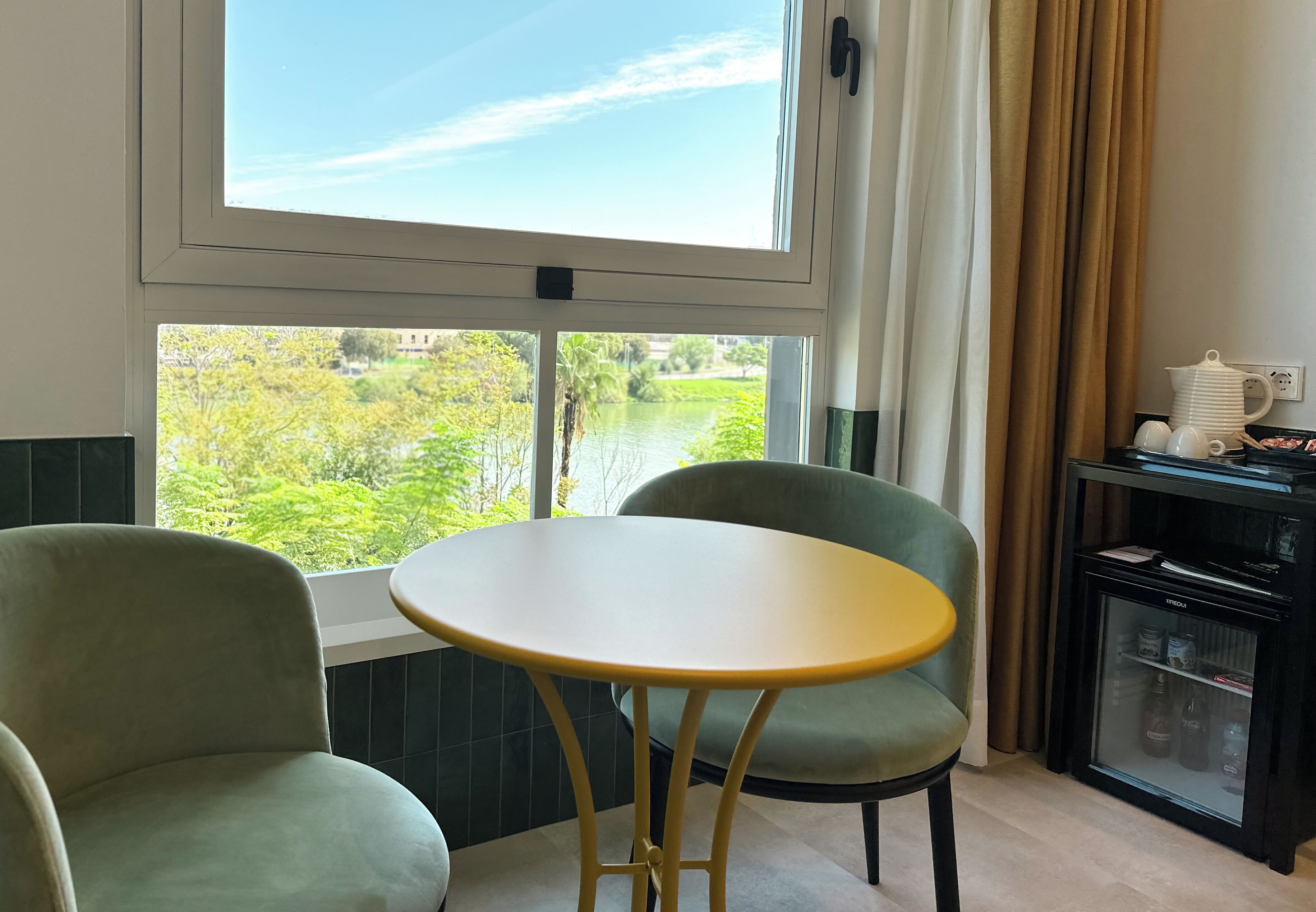 Una habitación con vista a vegetación y un río, una pequeña mesa redonda, dos sillas y un mini refrigerador con botellas.