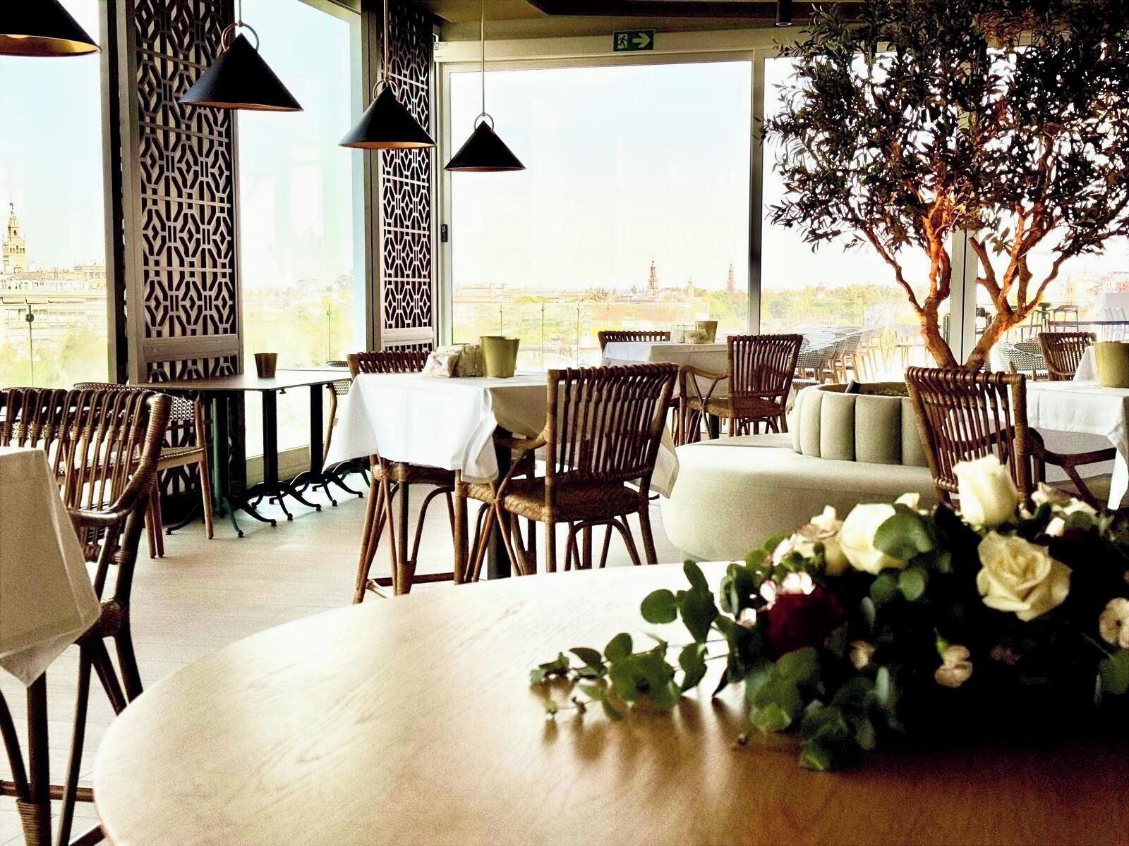 Un restaurante con mesas altas, sillas de mimbre, un árbol grande y vistas a un paisaje urbano.