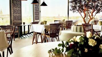Un restaurante con mesas altas, sillas de mimbre, un árbol grande y vistas a un paisaje urbano.