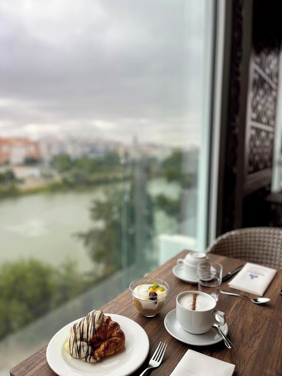 A table with a croissant, a dessert bowl, a cup of coffee, and a view of a river and cityscape.