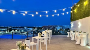 Una terraza al aire libre con mesas y sillas blancas, luces colgantes y vistas a un paisaje urbano al anochecer.