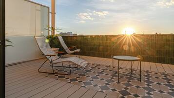 Un balcón con dos sillones reclinables a rayas y una pequeña mesa redonda, con vista a un paisaje urbano y la puesta de sol a lo lejos.