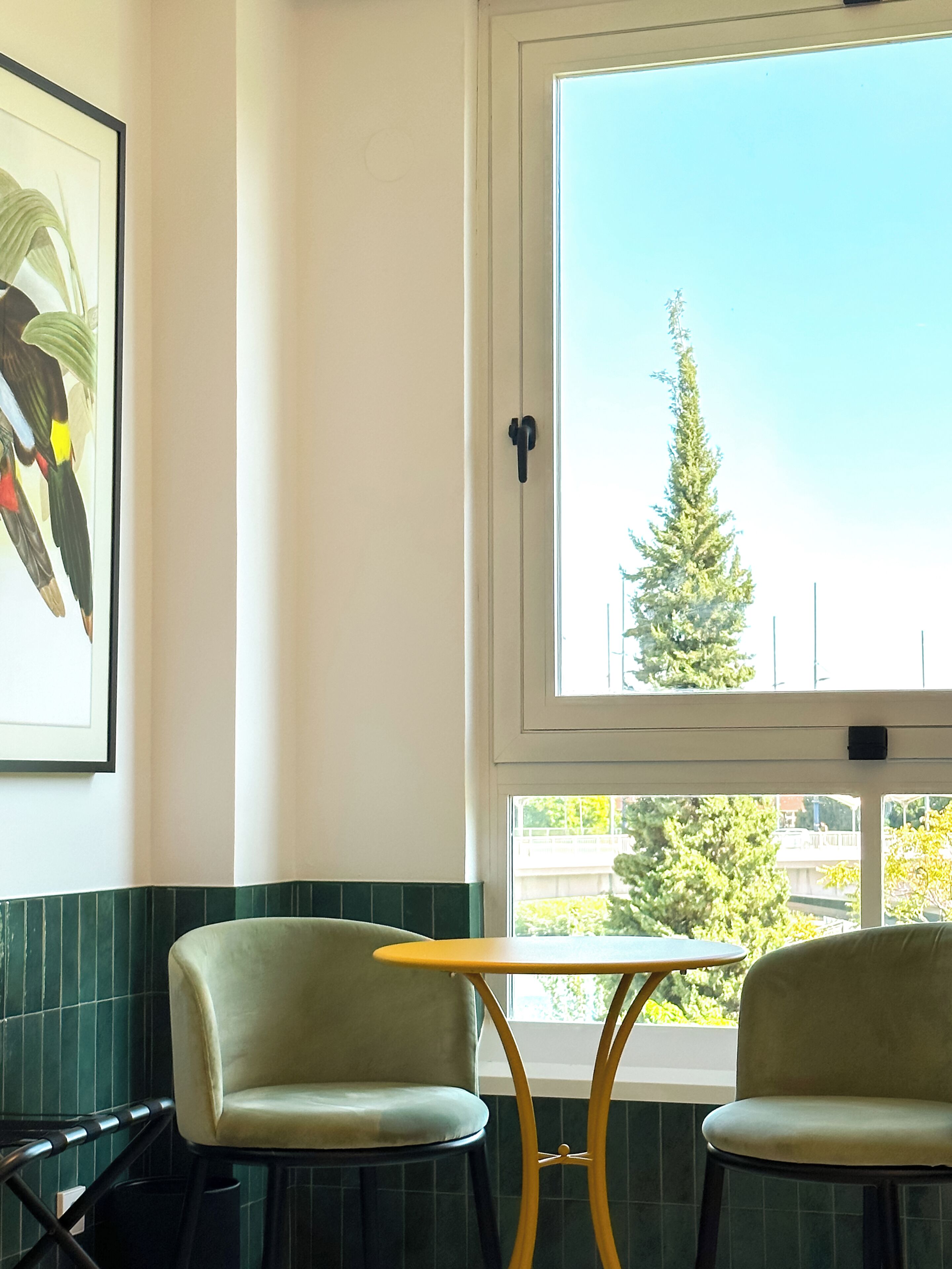 A room with two chairs, a small table, a window with a view of a tree, and a framed picture on the wall.