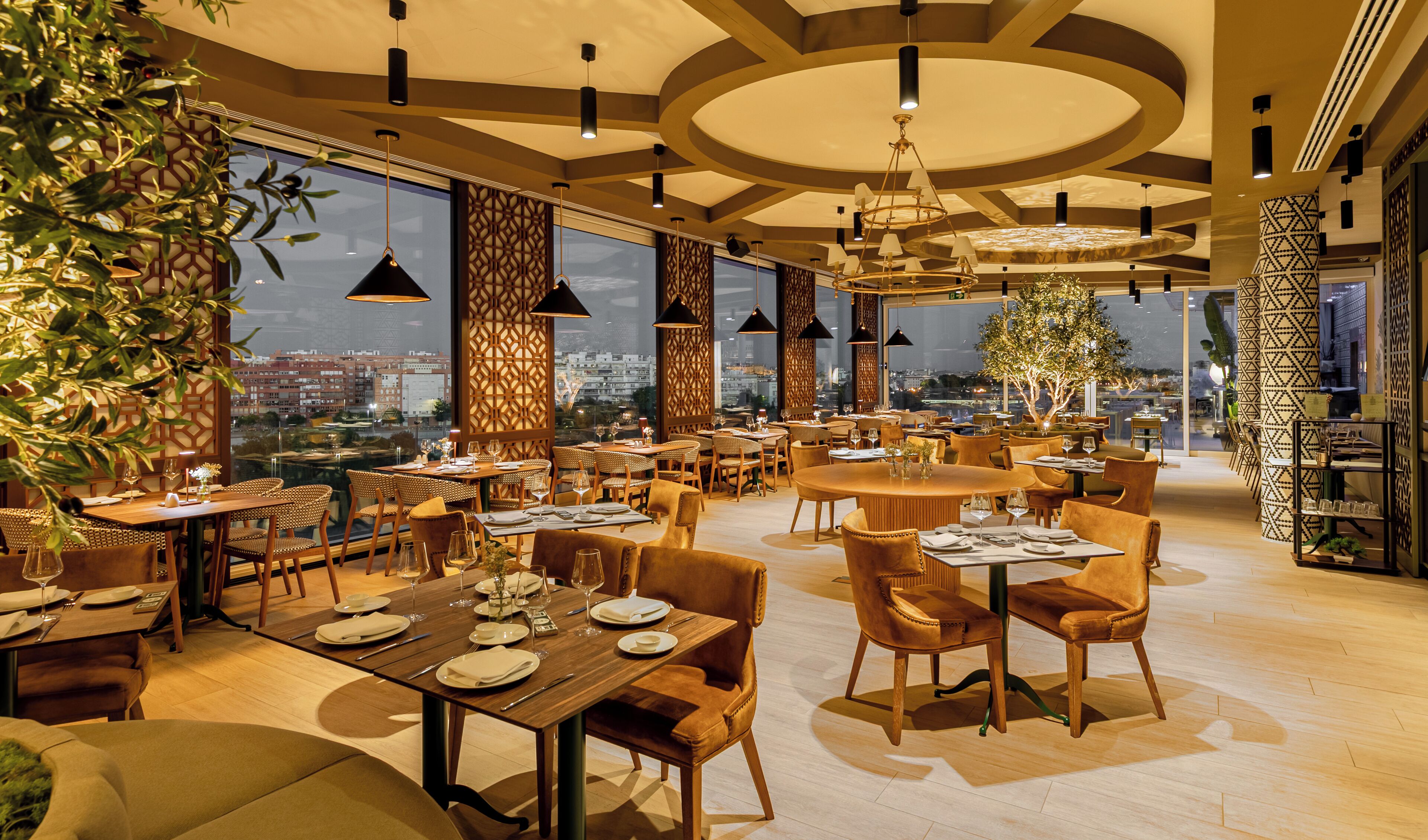 A modern restaurant with high ceilings, large windows, and a city view. Tables are set with white tablecloths and wine glasses.