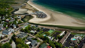 Vista aérea de un pueblo costero con playa, un puerto deportivo y un complejo hotelero grande con varios edificios y zonas de estacionamiento.