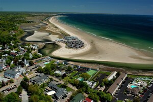 An aerial view of a coastal town with a beach, a marina, and a large hotel complex with multiple buildings and parking areas.