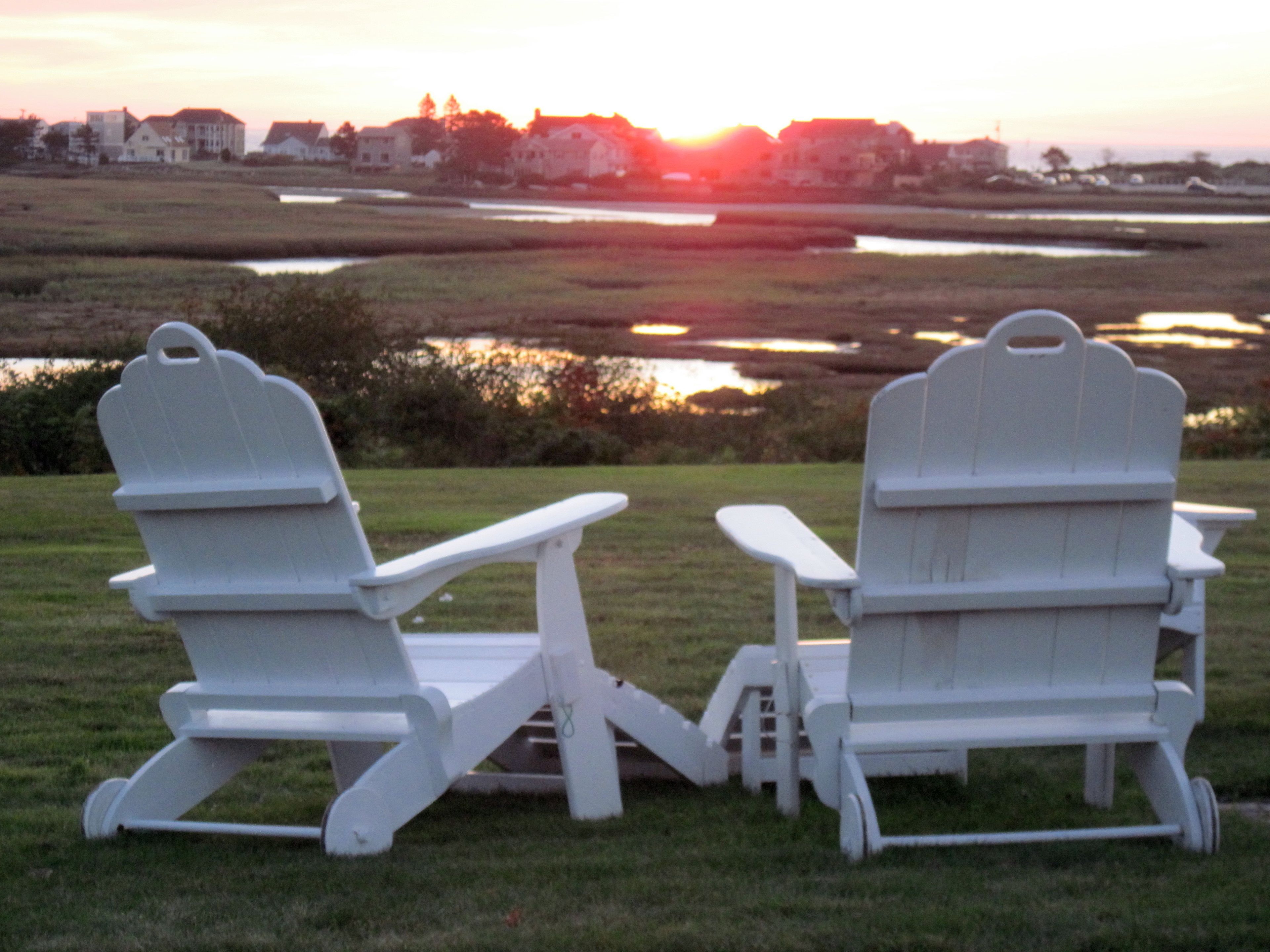 Two white Adirondack chairs facing each other with a small table in between, set on a grassy area with a view of a sunset over a body of water and buildings in the distance.