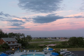 A swimming pool area with lounge chairs and umbrellas, a small building with a sign, and a view of a golf course and houses in the background. - The Mariner Resort (Ogunquit)
