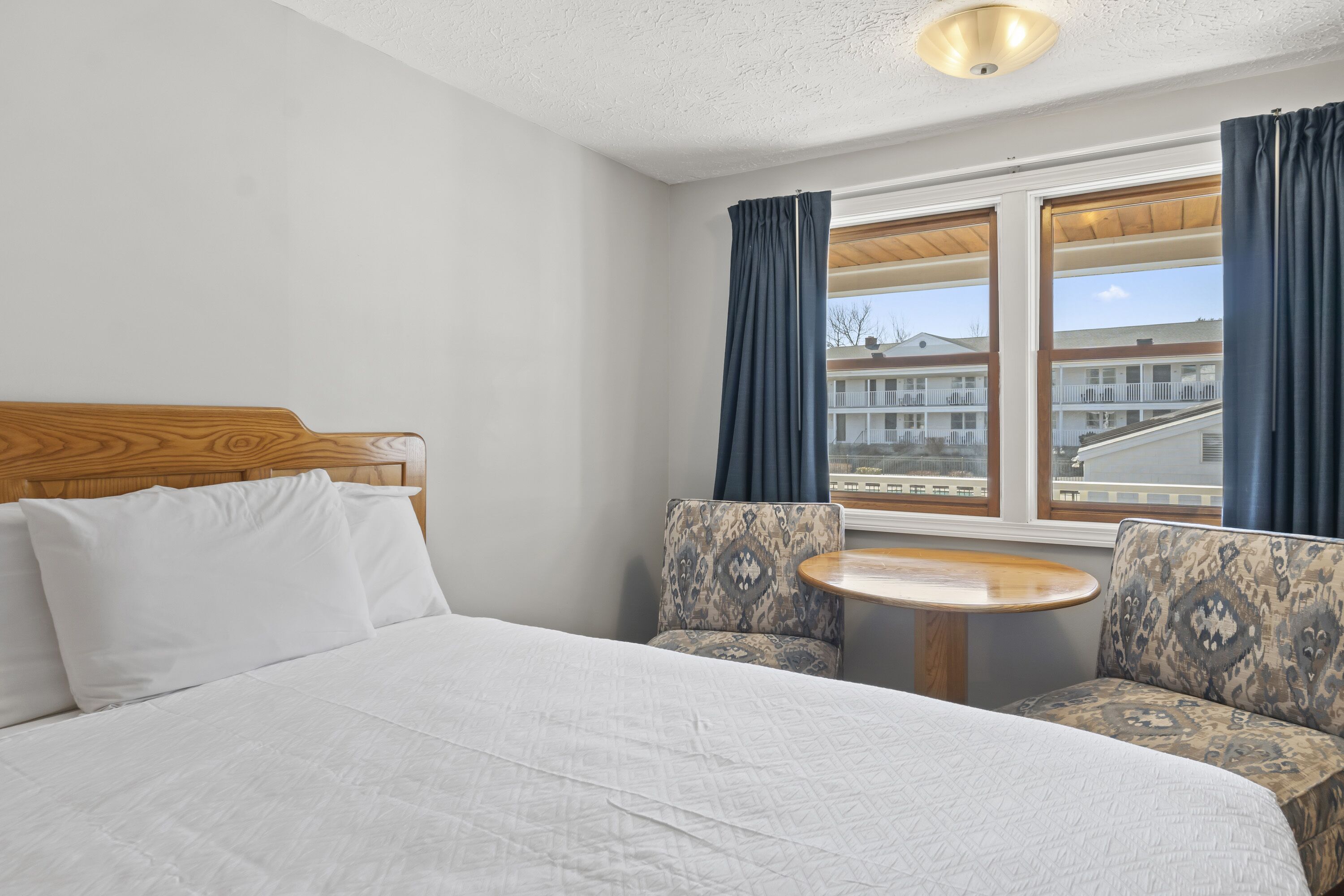 Une chambre d’hôtel avec un lit, deux fauteuils à motifs, une petite table ronde et une fenêtre avec des rideaux.