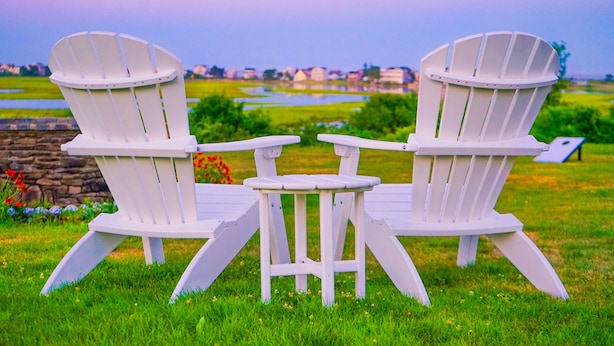 Dos sillas Adirondack blancas y una mesita instaladas en un campo de césped, con vista a un lago y edificios a lo lejos.