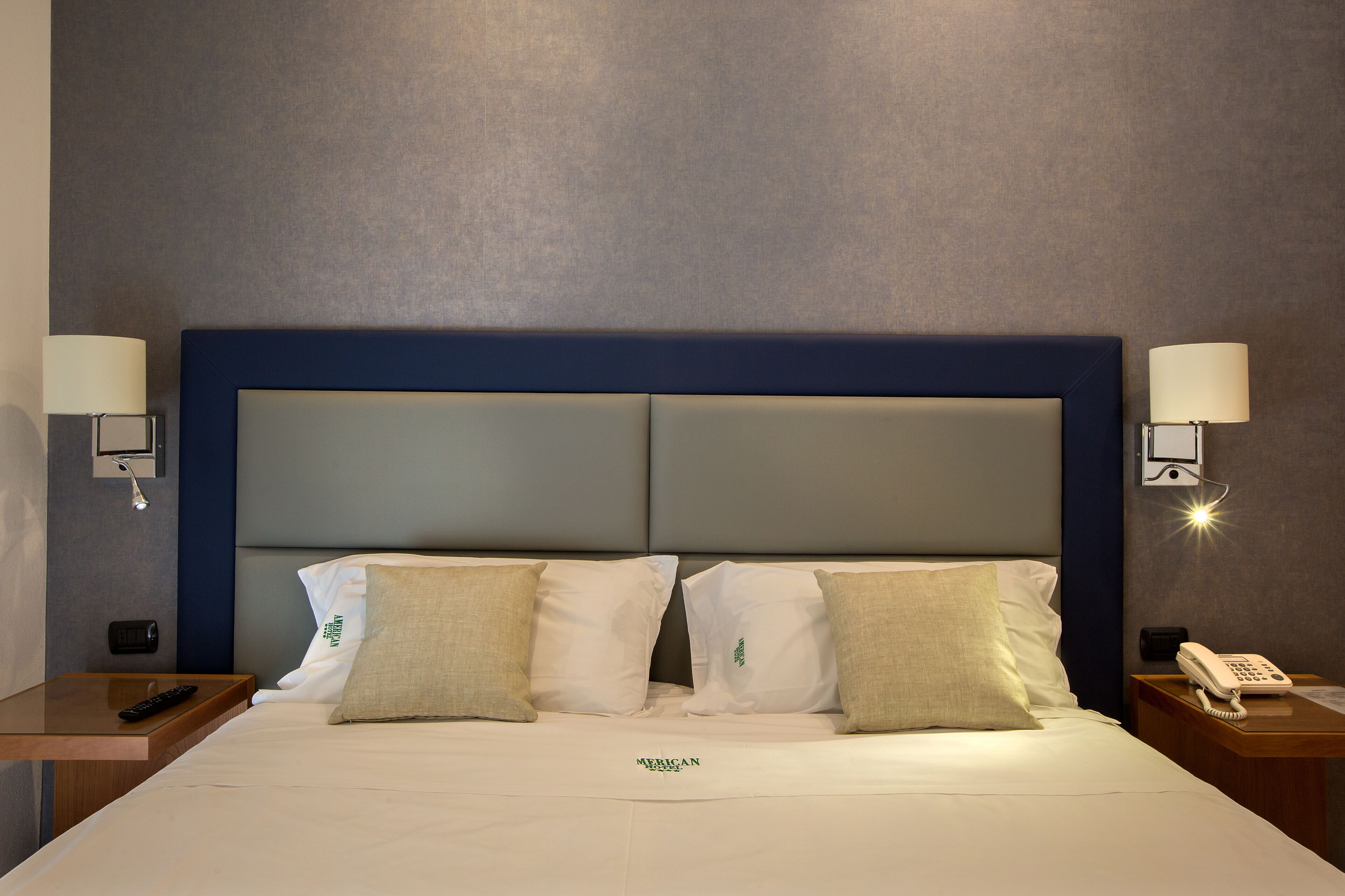 Een hotelkamer met een groot bed, twee nachtkastjes, een telefoon aan de muur en een lamp aan beide zijden.