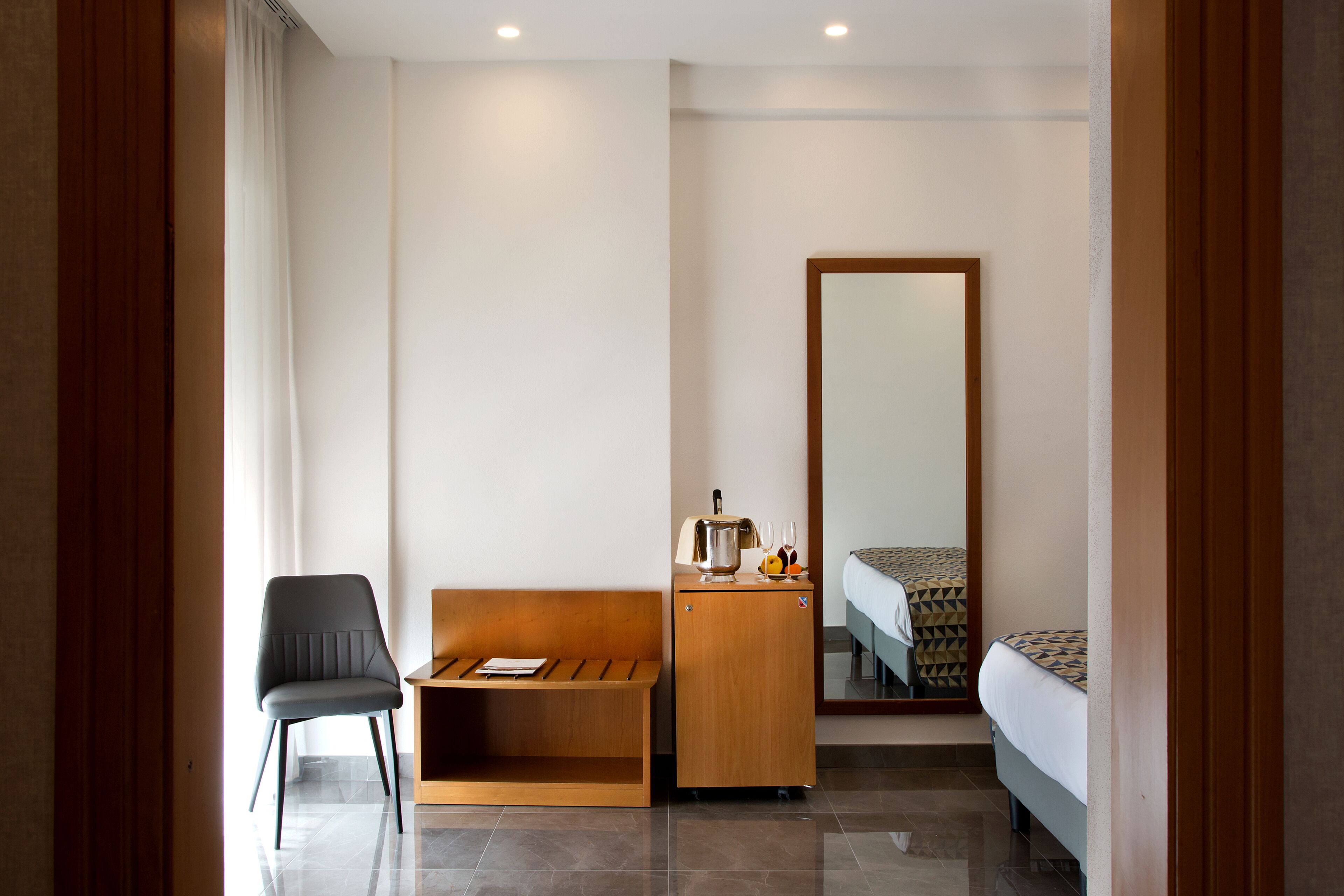A hotel room with a wooden bench, a small wooden cabinet, a bed with patterned bedding, and a large mirror on the wall.