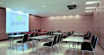 A conference room with a projection screen displaying the American hotel logo, multiple tables with chairs, and a podium.