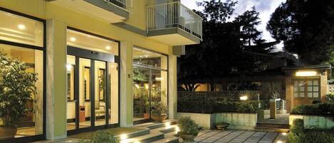 A multi-story hotel with a well-lit entrance, outdoor seating area, and a landscaped garden.