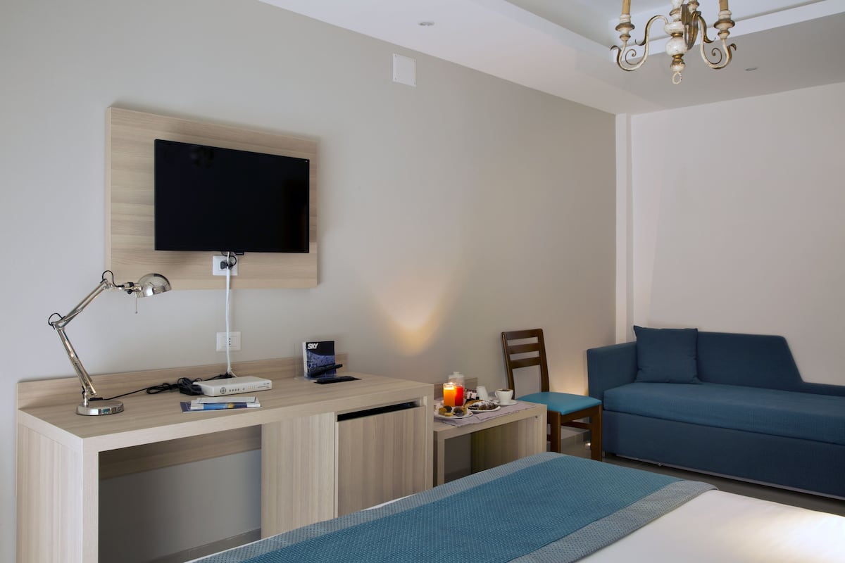 A hotel room with a bed, a desk with a computer, a TV, a chair, and a blue sofa.