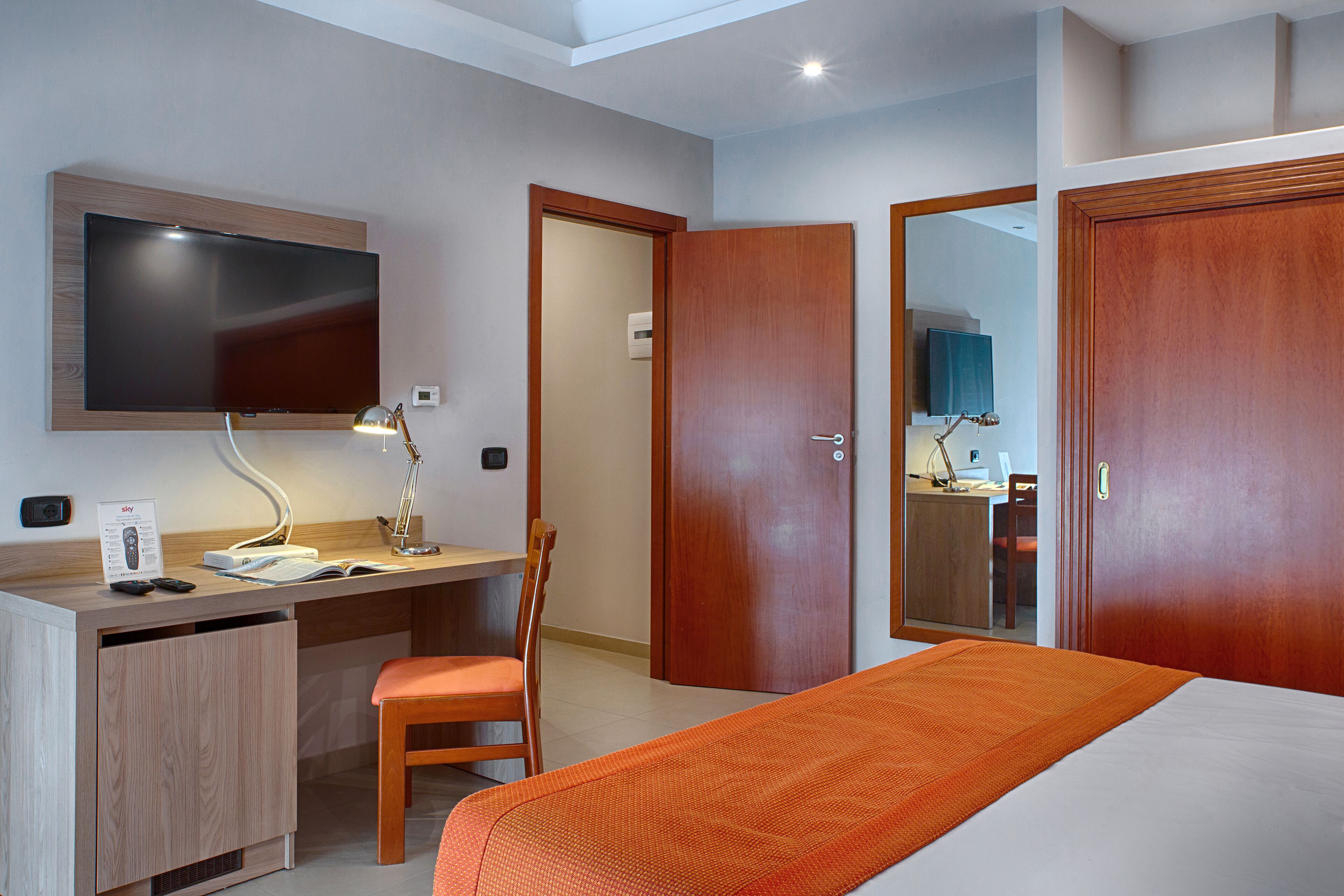 Ein Hotelzimmer mit einem Bett, einem Schreibtisch mit Computer, einem Sessel, einem Fernseher und zwei Türen.