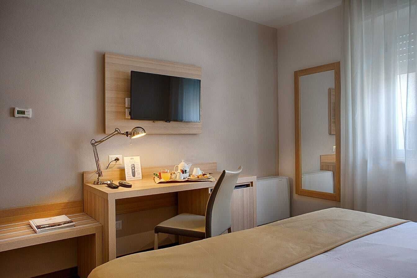 Habitación de hotel con cama, escritorio, silla, televisor, espejo y ventana con cortinas.