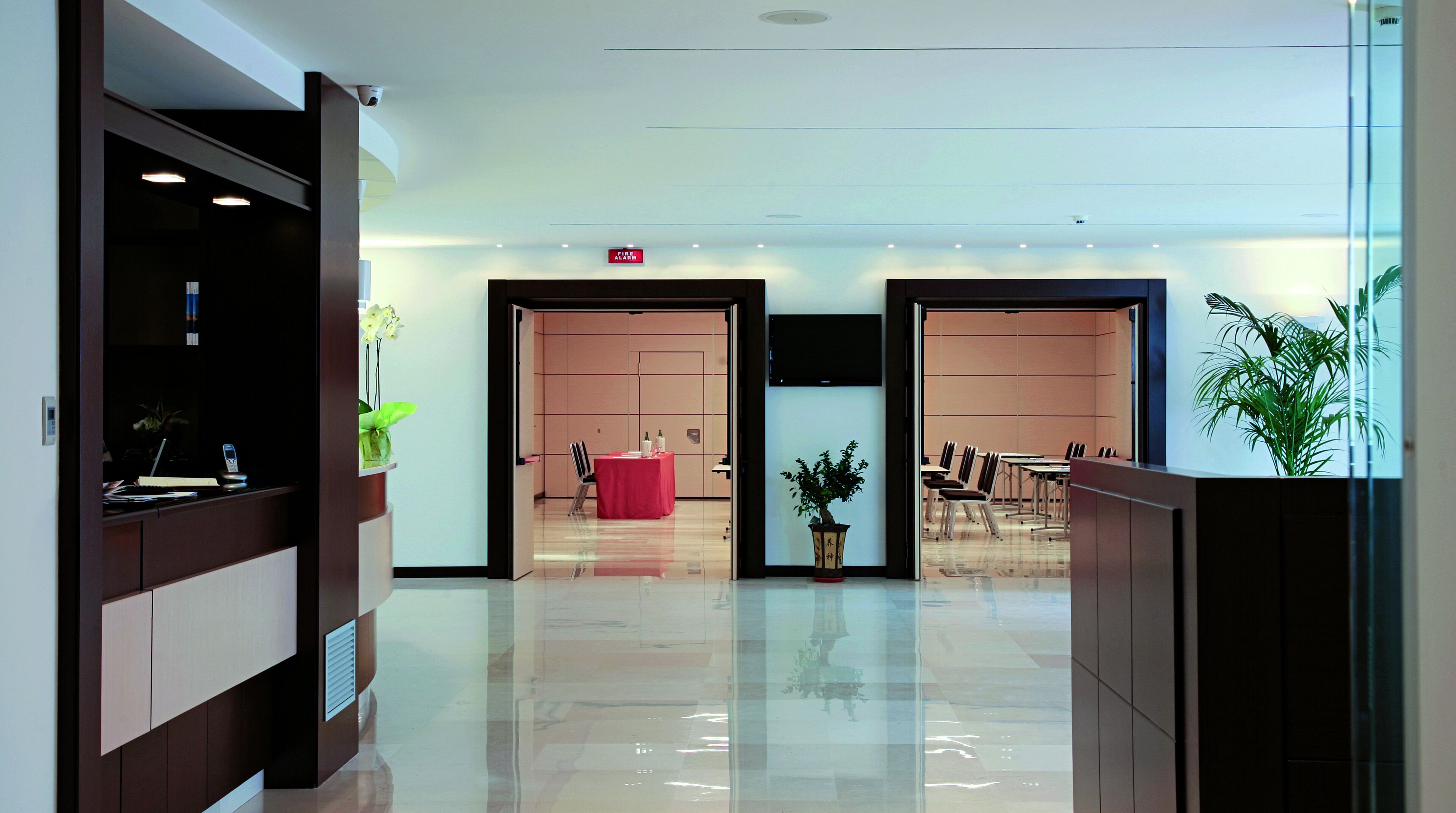 A modern hotel lobby with a reception desk, a red seating area, and a conference room with chairs and tables.