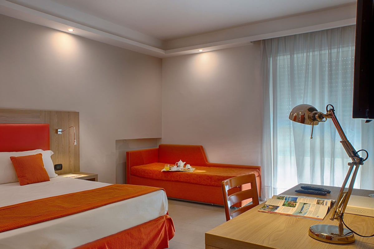 A hotel room with a bed, a sofa, a desk with a lamp, and a television.