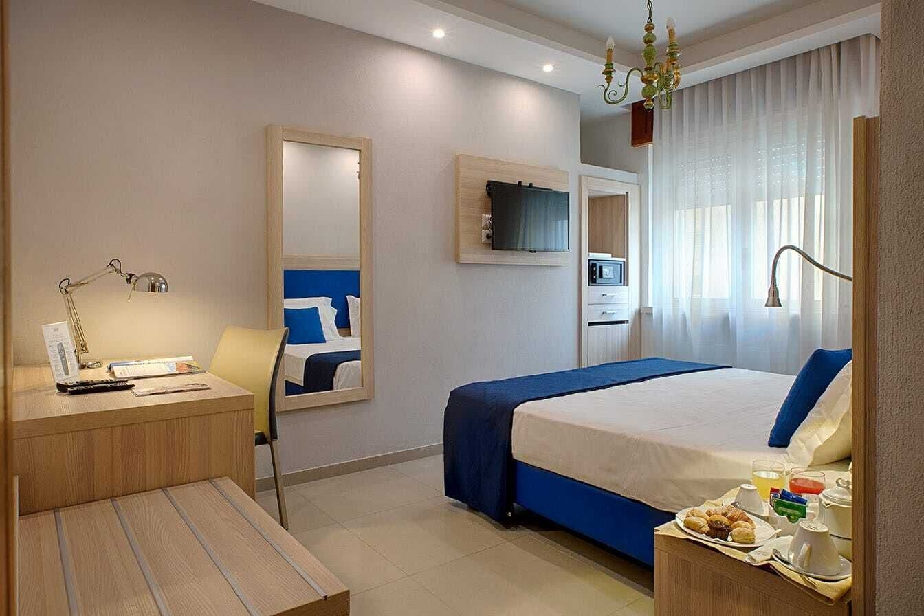 Una camera d'albergo con un letto, una scrivania con una lampada, una sedia, una televisione e un vassoio per la colazione.