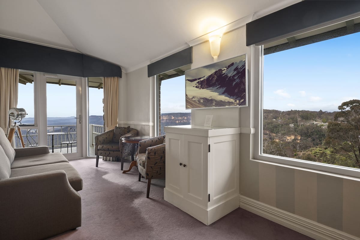 A room with a balcony, a sofa, two patterned armchairs, a cabinet, and a painting of a mountain landscape.