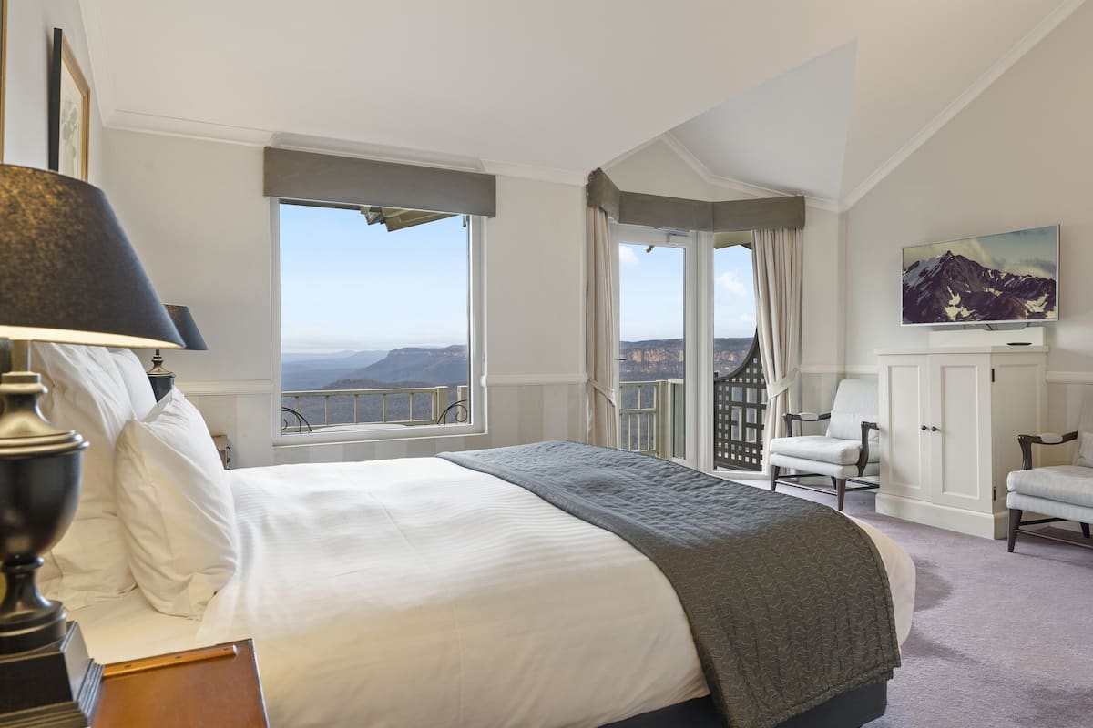A bedroom with a large bed, a view of mountains, a balcony with a chair, and a painting on the wall.