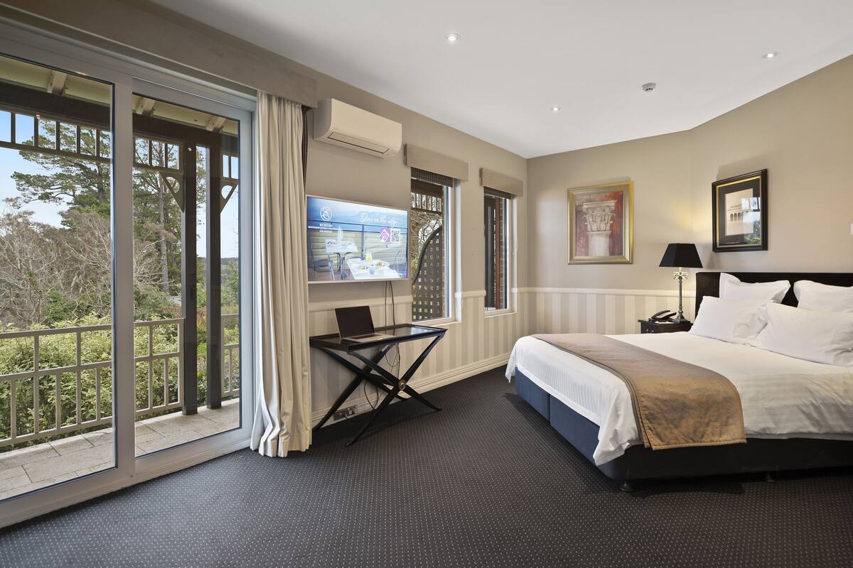 A hotel room with a large bed, a desk with a laptop, a TV, and a balcony with a view of trees.