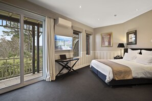 A hotel room with a large bed, a desk with a laptop, a TV, and a balcony with a view of trees.