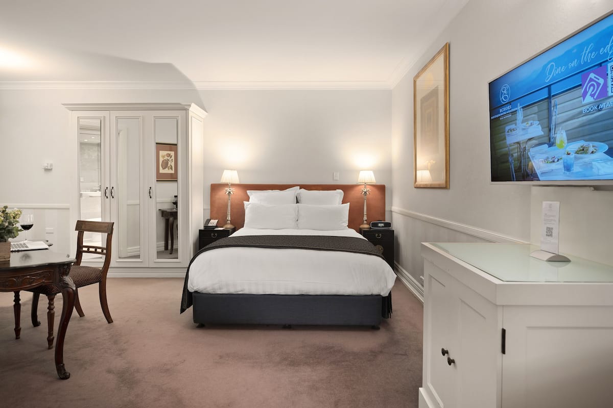 A hotel room with a bed, bedside tables, a desk with a chair, a wardrobe, and a TV on the wall.