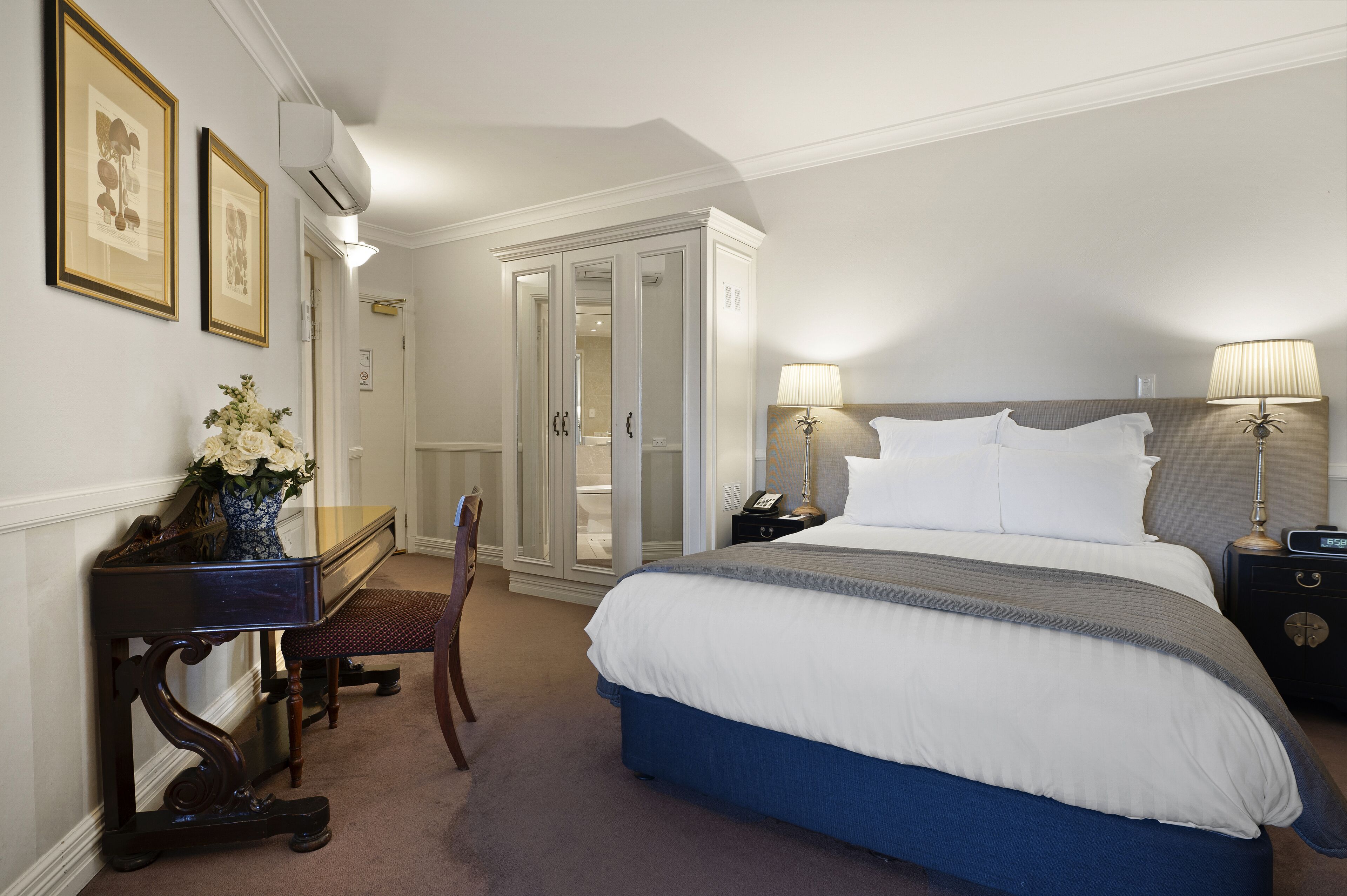 A hotel room with a large bed, bedside lamps, a desk with a vase of flowers, and a wardrobe.