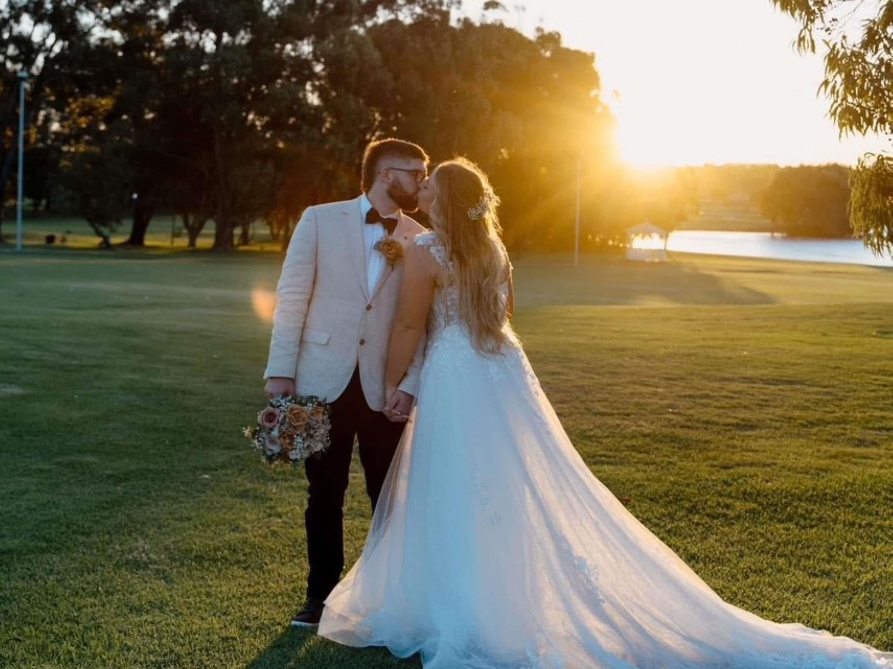 A couple in wedding attire on a grassy field with a body of water and trees in the background.