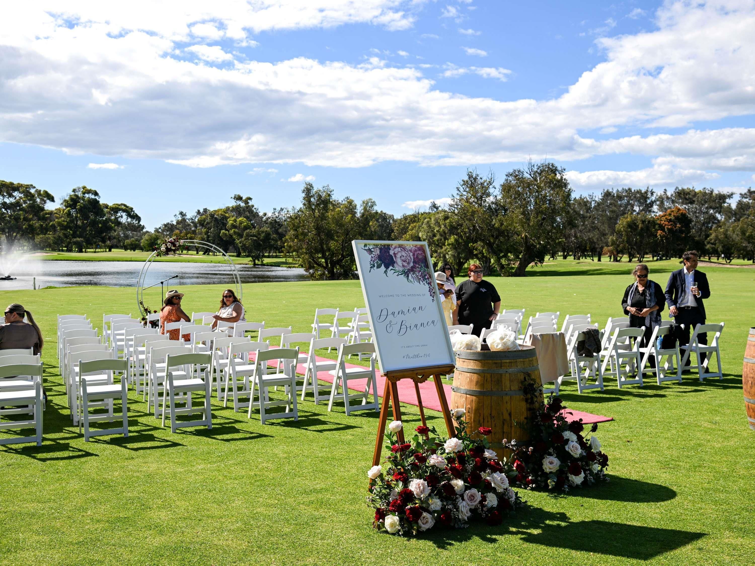 A wedding ceremony setup with white chairs, a wooden barrel, and floral arrangements on a grassy field.
