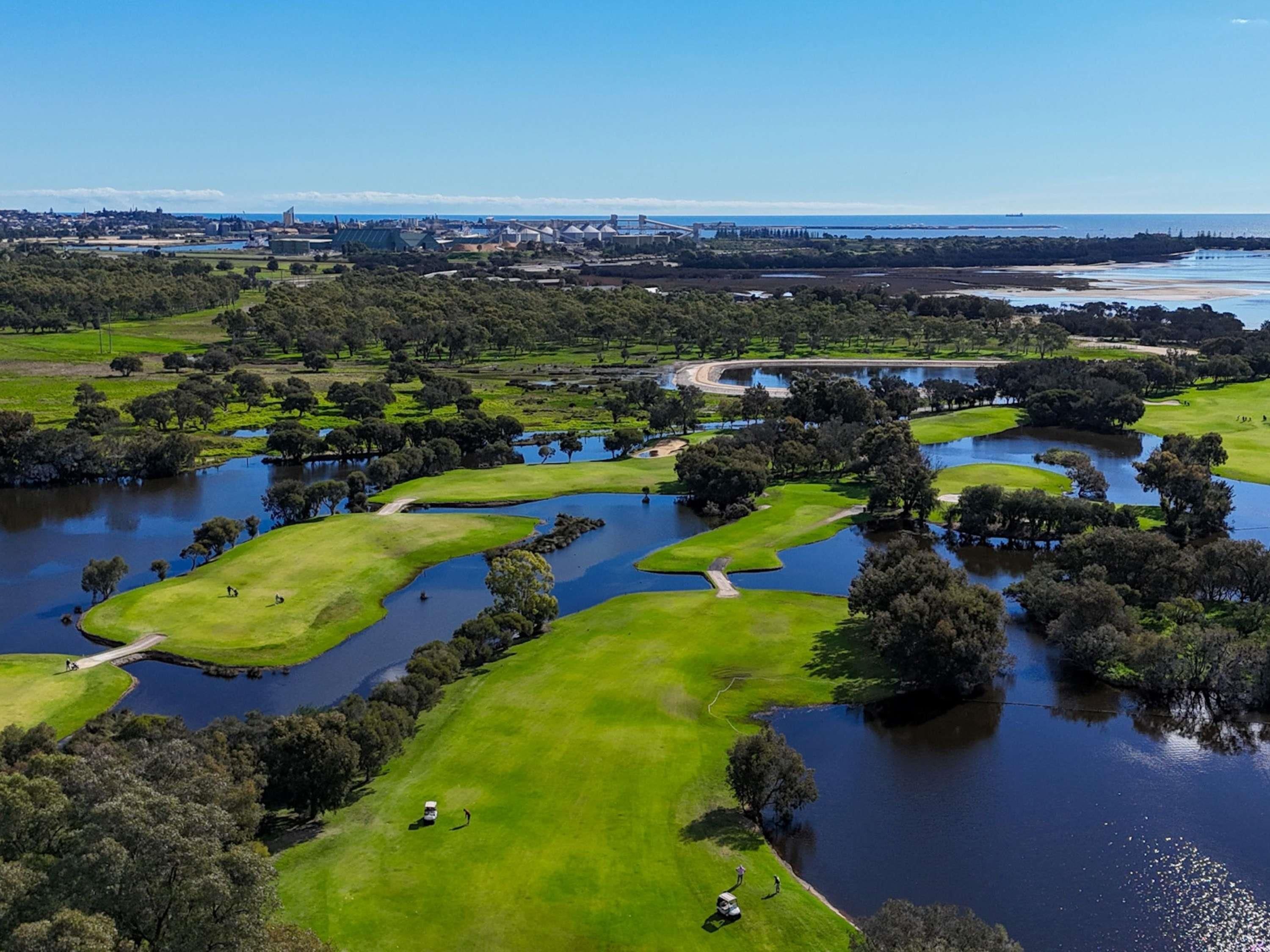 An aerial view of a golf course with water hazards, surrounded by greenery and a distant cityscape.