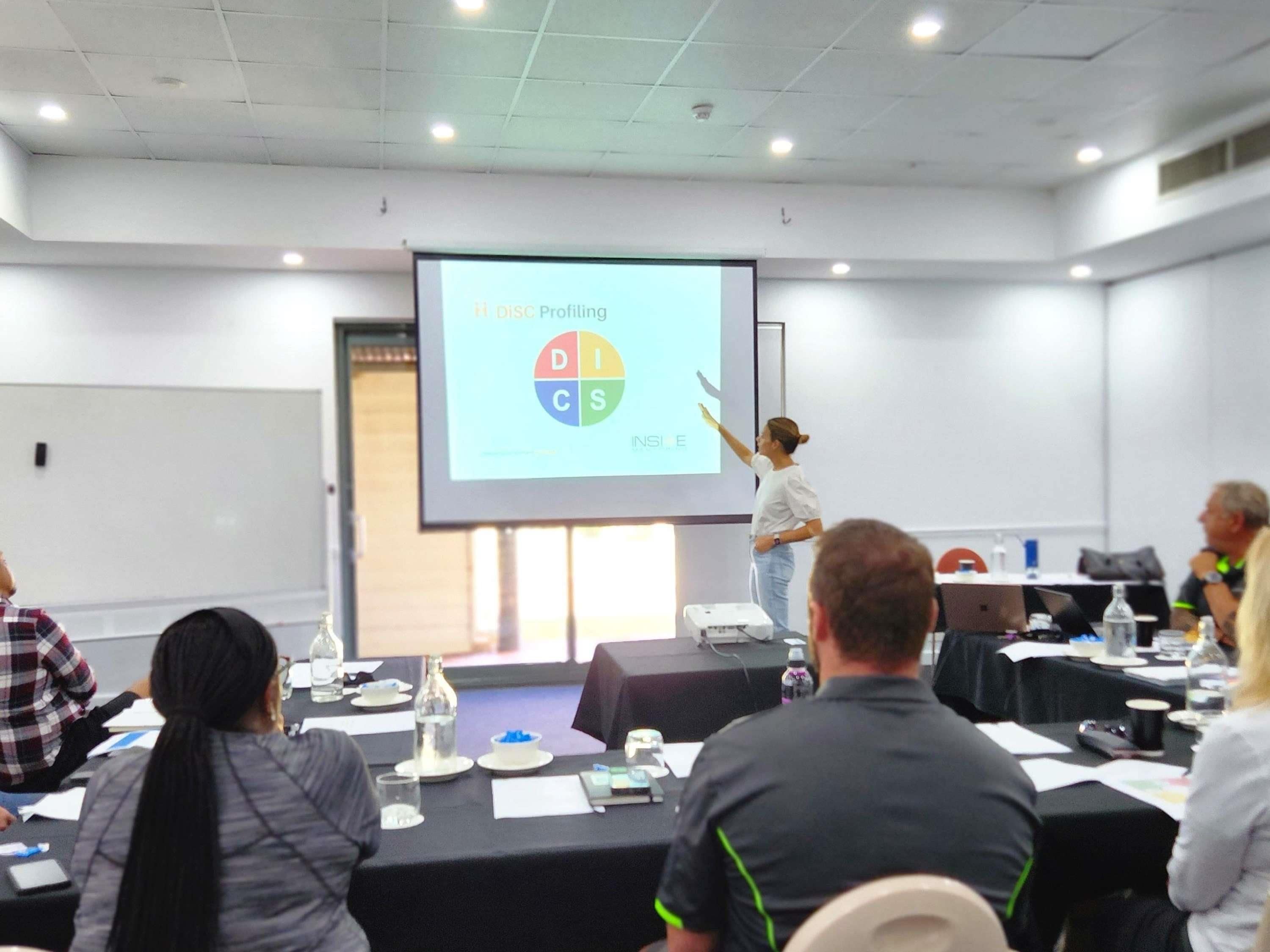 A conference room with a presentation screen displaying 'DISC Profiling INSIDE,' a projector, and attendees seated at tables with water bottles and papers.