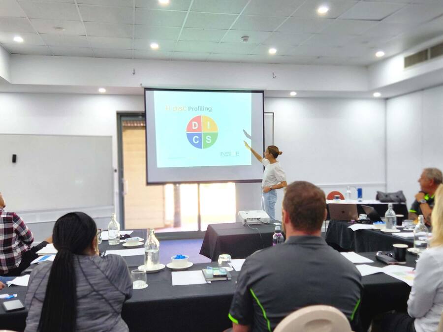 Un salón de conferencias con una pantalla mostrando 'DISC Profiling INSIDE', un proyector, y asistentes sentados en mesas con botellas de agua y documentos.