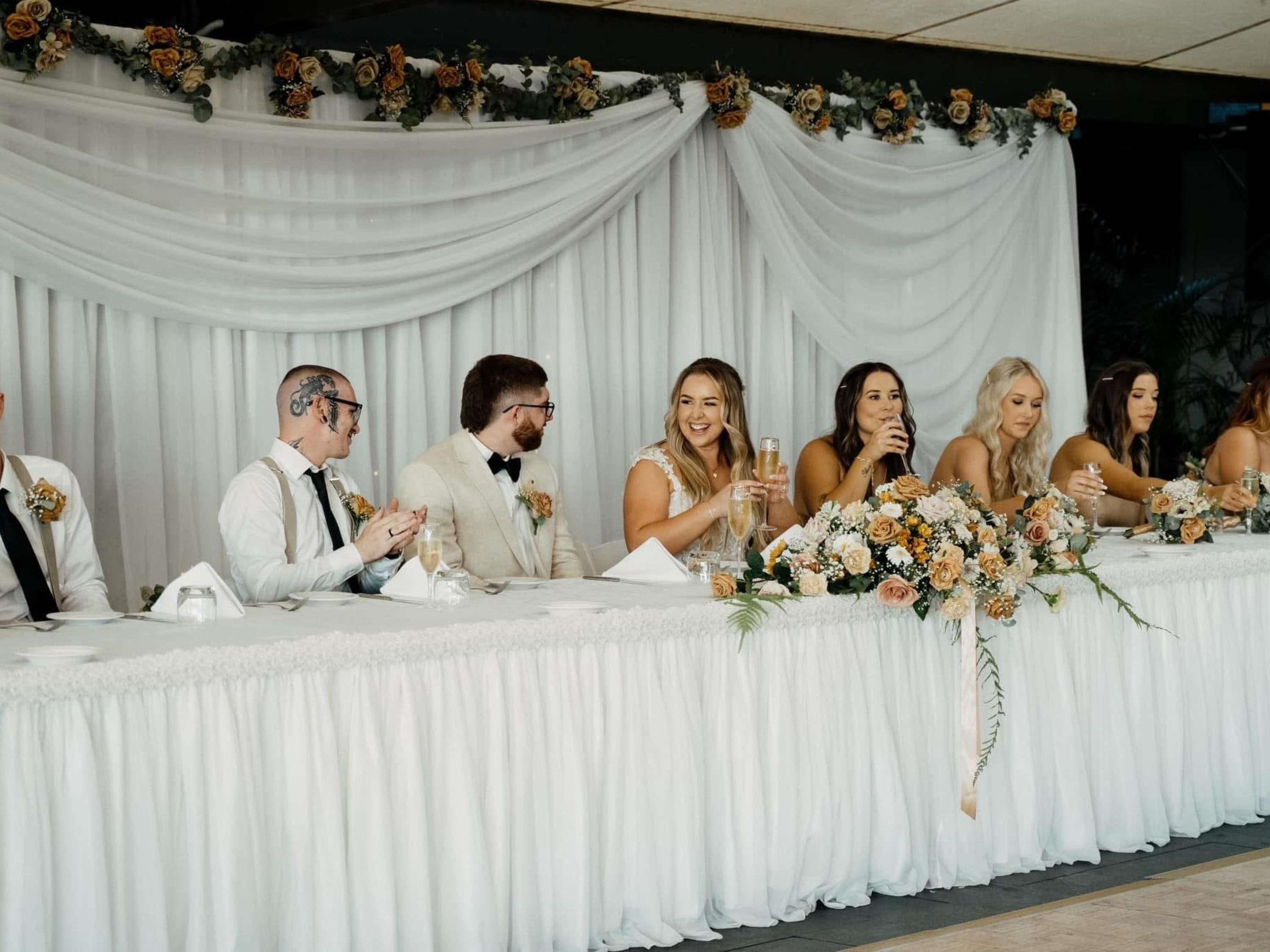 A wedding reception with a white tablecloth, floral arrangements, and guests seated at a long table.