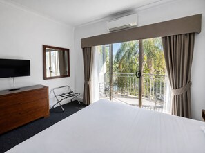 Habitación de hotel con una cama, un televisor sobre un tocador de madera, un balcón con cortinas y vistas a palmeras.