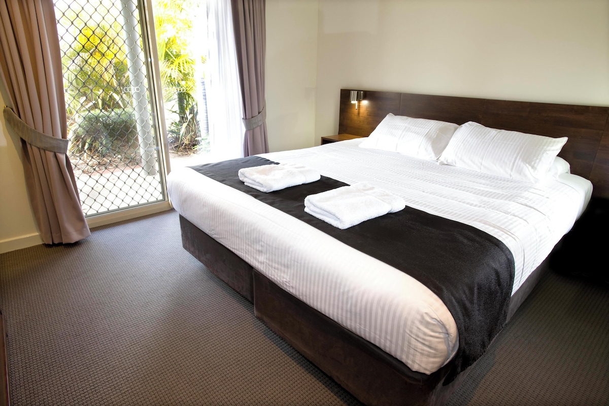 Ein Hotelzimmer mit einem großen Bett, einem Kopfteil mit einer Leuchte und Blick auf Grün durch ein Fenster mit Vorhängen.