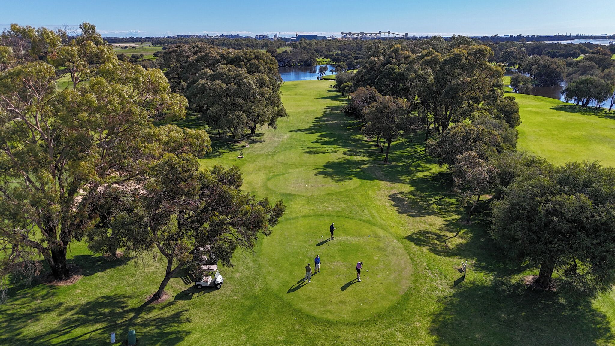 An aerial view of a golf course with trees, a lake, and a bridge in the background.