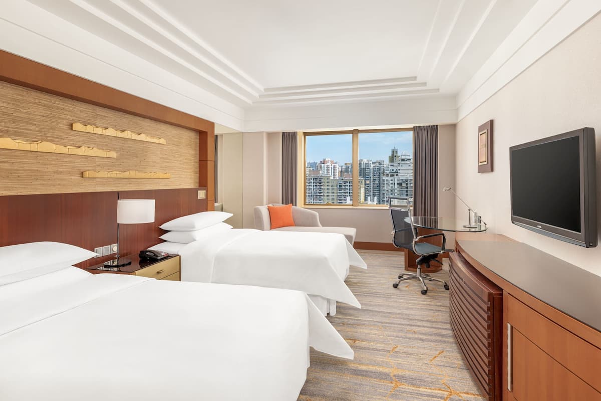 A hotel room with two beds, a desk, a TV, and a city view.