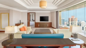 Habitación de hotel moderna con un ventanal grande que ofrece vistas a la ciudad, un televisor de pantalla plana montado en la pared, un sofá, una mesa de centro y una mesa de comedor con sillas.