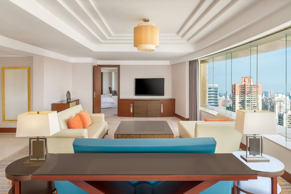 A modern hotel room with a large window offering a city view, a flat-screen TV mounted on the wall, a sofa, a coffee table, and a dining table with chairs.