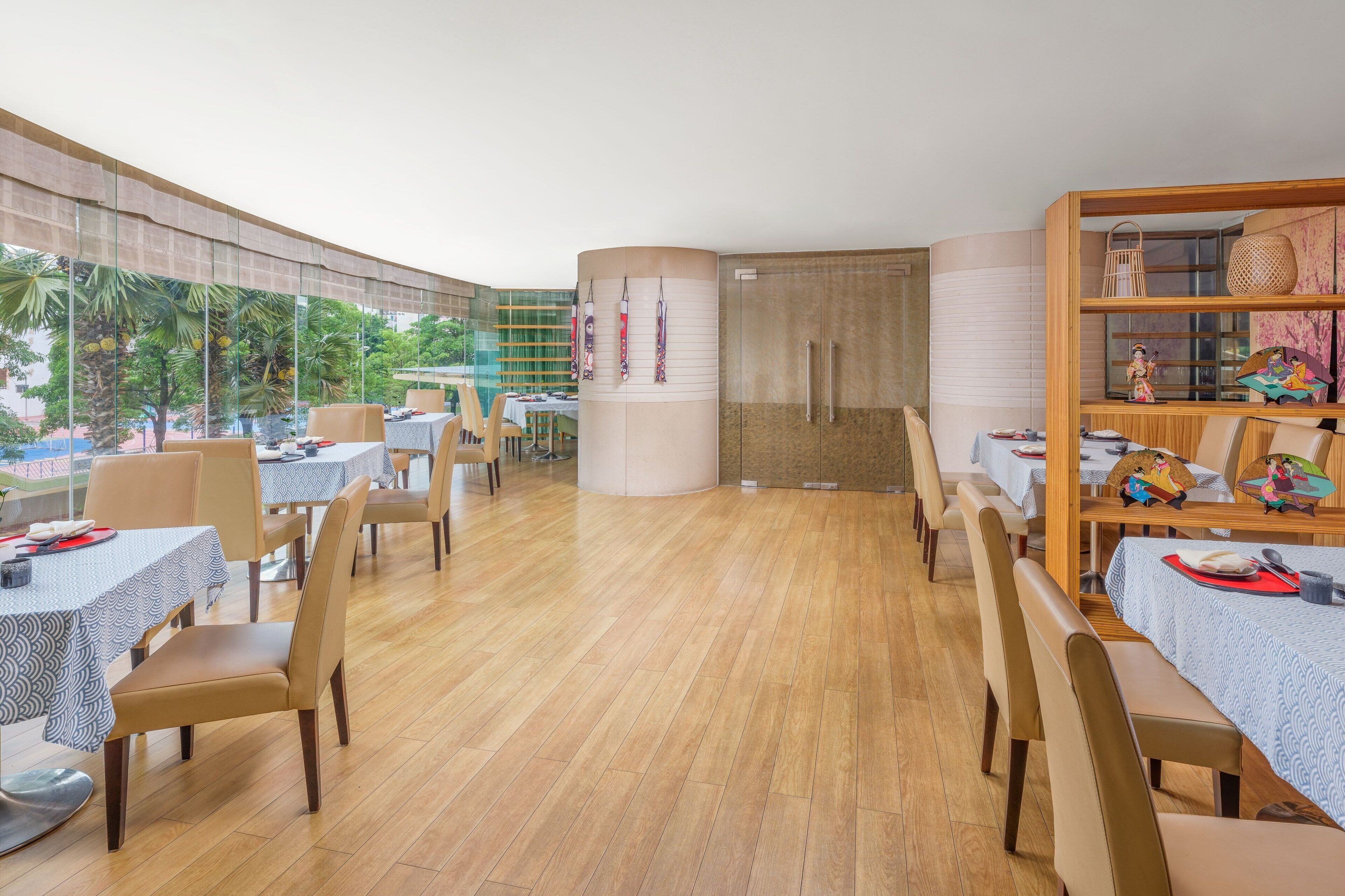 A spacious restaurant with wooden flooring, round tables, and chairs. There are large windows offering a view of palm trees and a building.