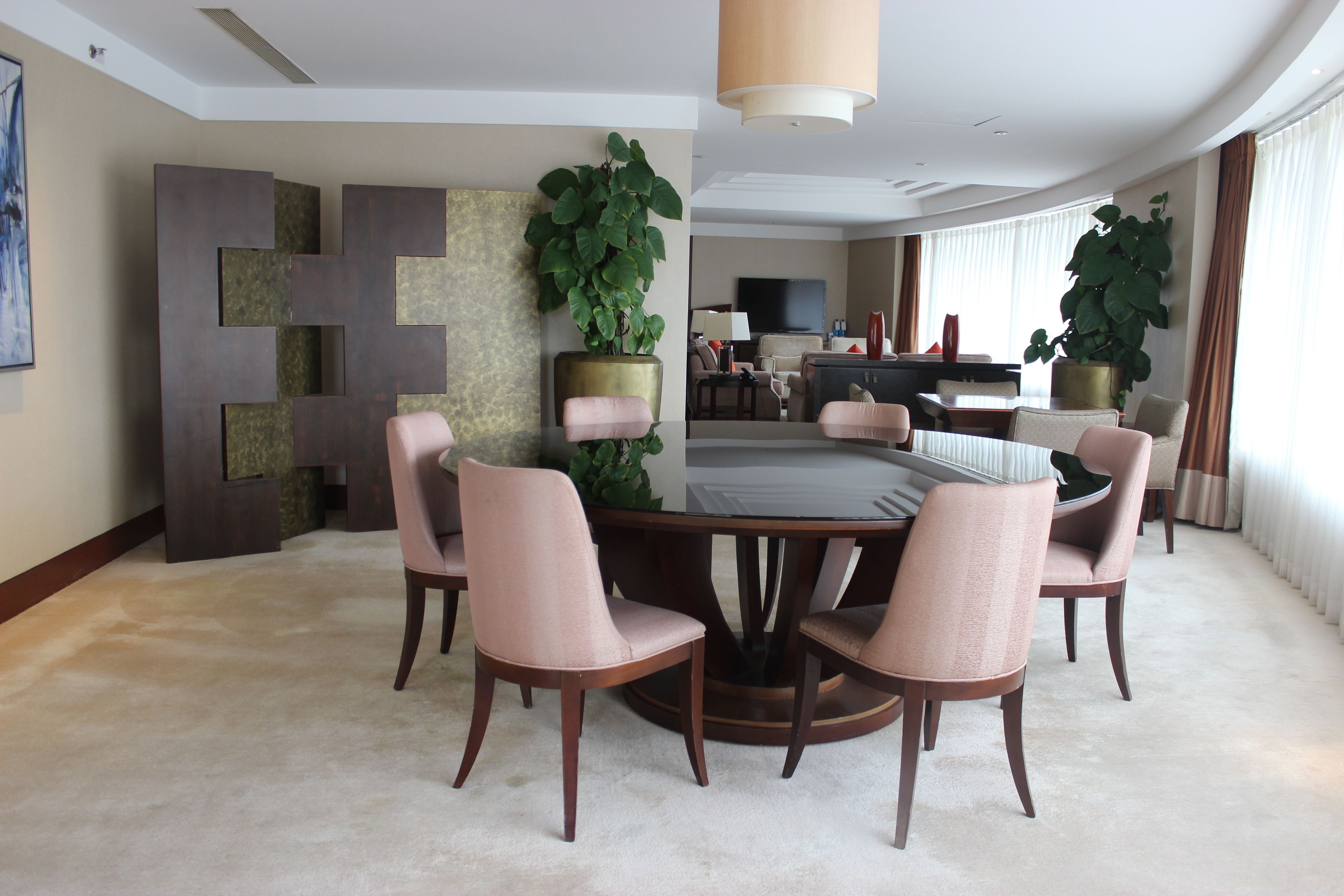 Un comedor moderno con una mesa redonda rodeada por seis sillas, una planta grande en maceta y un diseño contemporáneo en la pared.