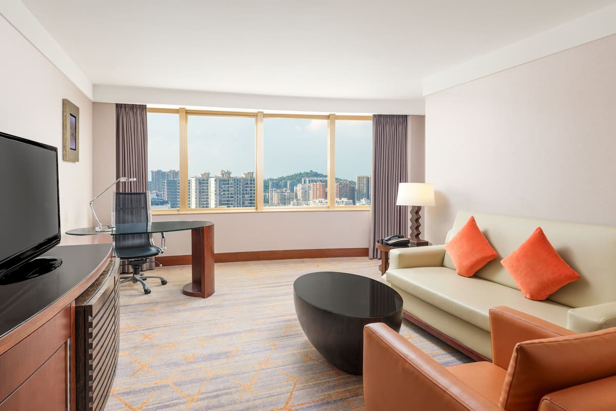A hotel room with a large window, a flat-screen TV, a sofa with orange cushions, a coffee table, a desk with a chair, and a lamp.